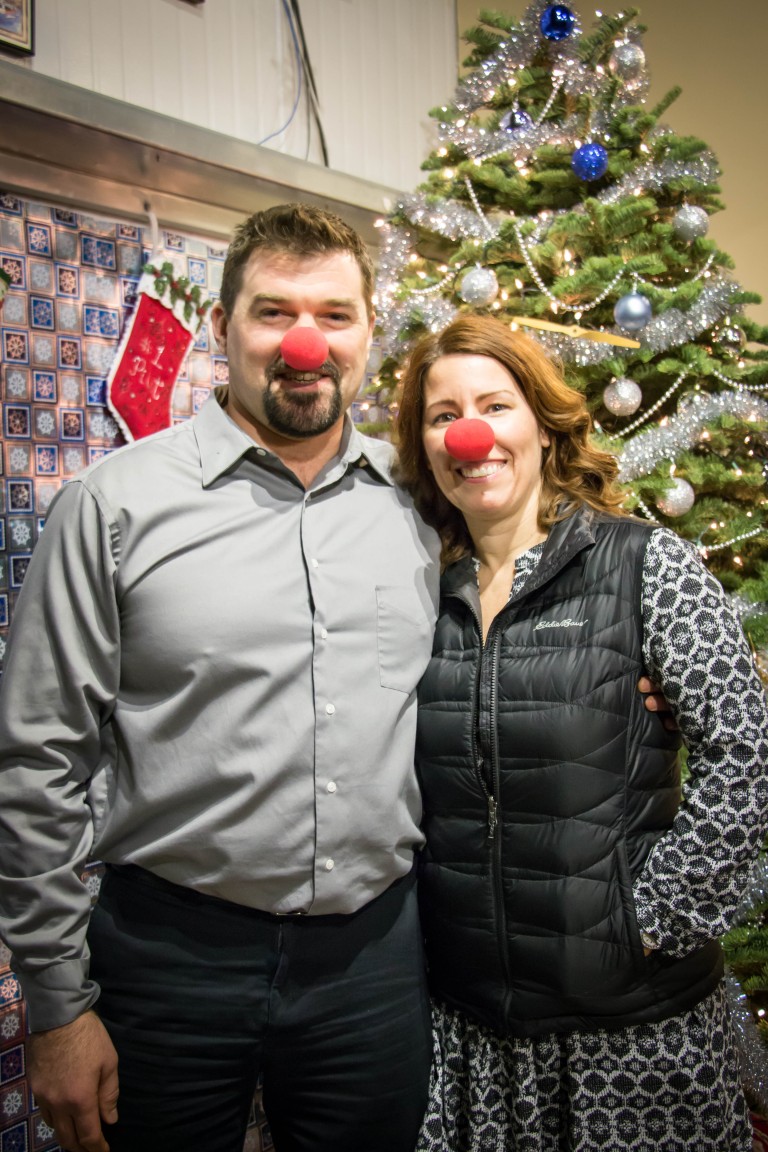 Aaron standing with a woman wearing red balls on their noses in front of a Christmas tree