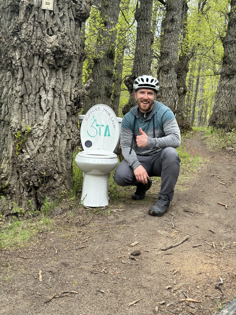 An AMC employee in a bikers helmet on a trail sitting next to a toilet