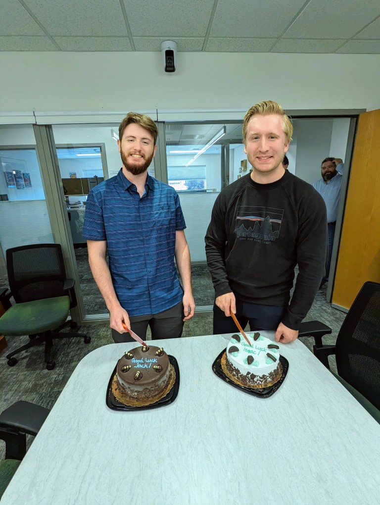 Two men posing in front of cakes that say Good Luck Jack and Good Luck Isaac
