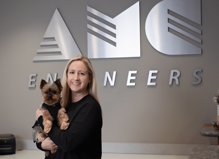 Woman holding yorkshire terrier in front of AMC Engineers logo panel on wall
